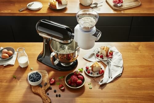 Kitchen mixer with ingredients and desserts on a wooden countertop.